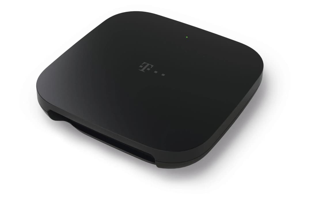 Ein schwarzes quadratisches Gerät mit dem T-Mobile-Logo oben. Ein grünes Licht ist sichtbar. Es steht auf einer weißen Oberfläche.